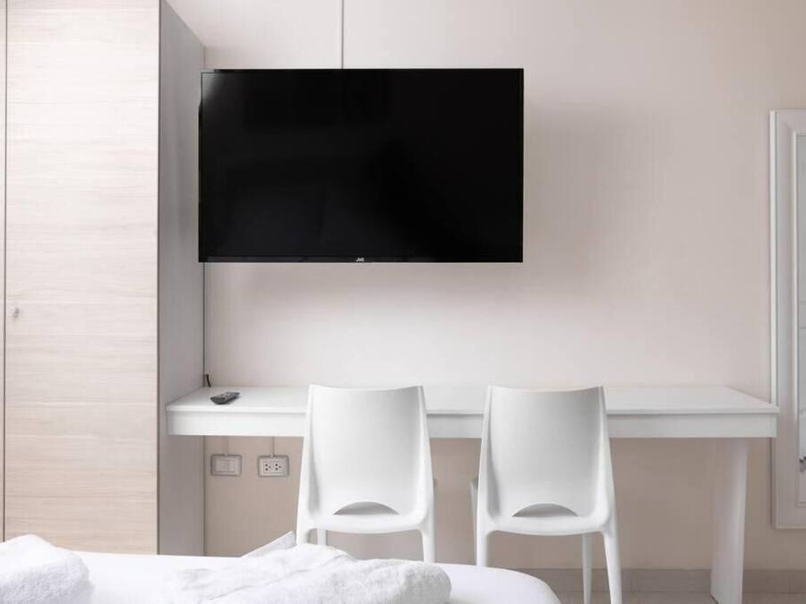 Una habitación de hotel moderna con una televisión de pantalla plana sobre un escritorio blanco, dos sillas blancas y una cama con una manta blanca.