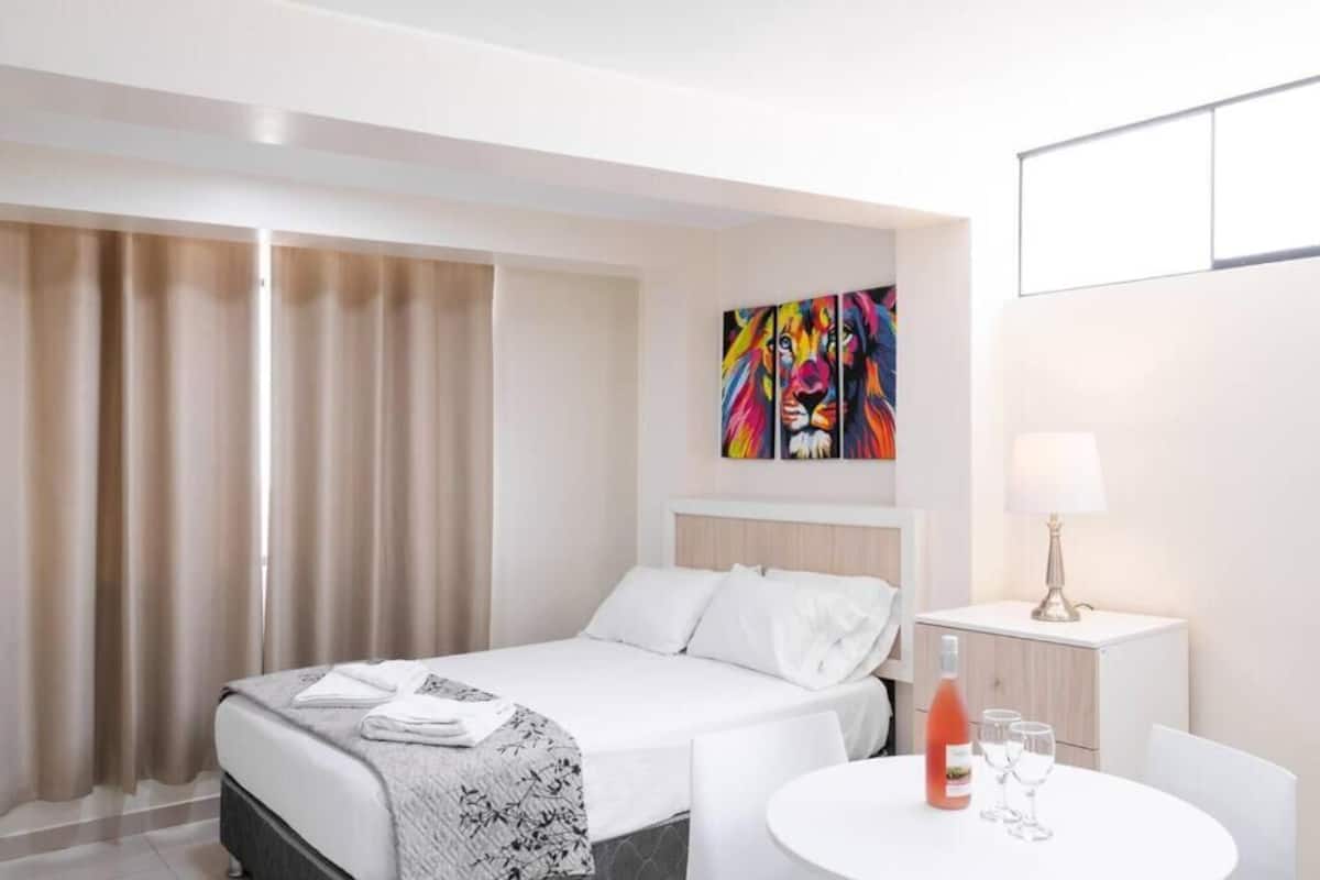 Ein Hotelzimmer mit einem Bett, einem Nachttisch mit Lampe, einem kleinen runden Tisch mit Flasche und Gläsern sowie einem farbenfrohen abstrakten Gemälde an der Wand.