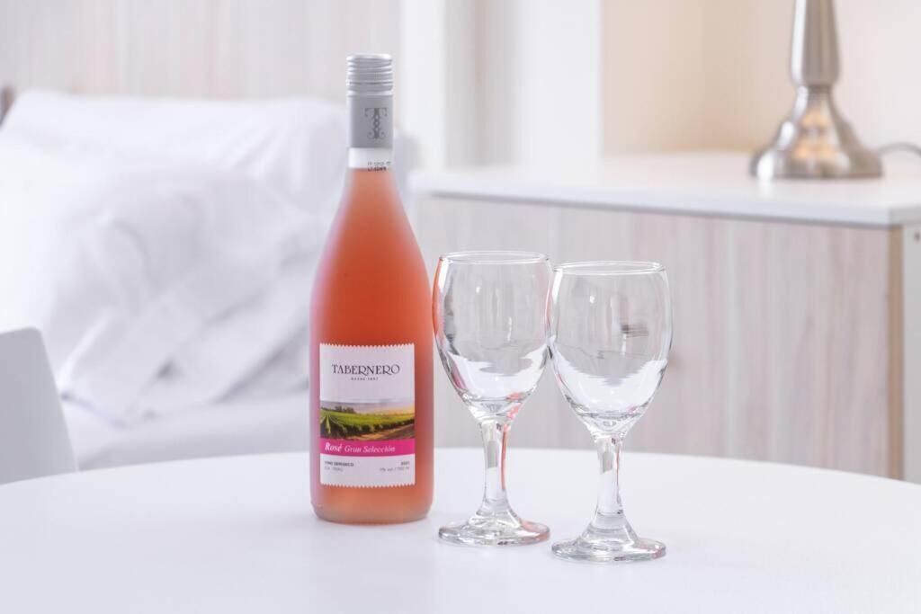 A bottle of Tabernero wine and two glasses on a white table in a room with a white bed and a lamp in the background.