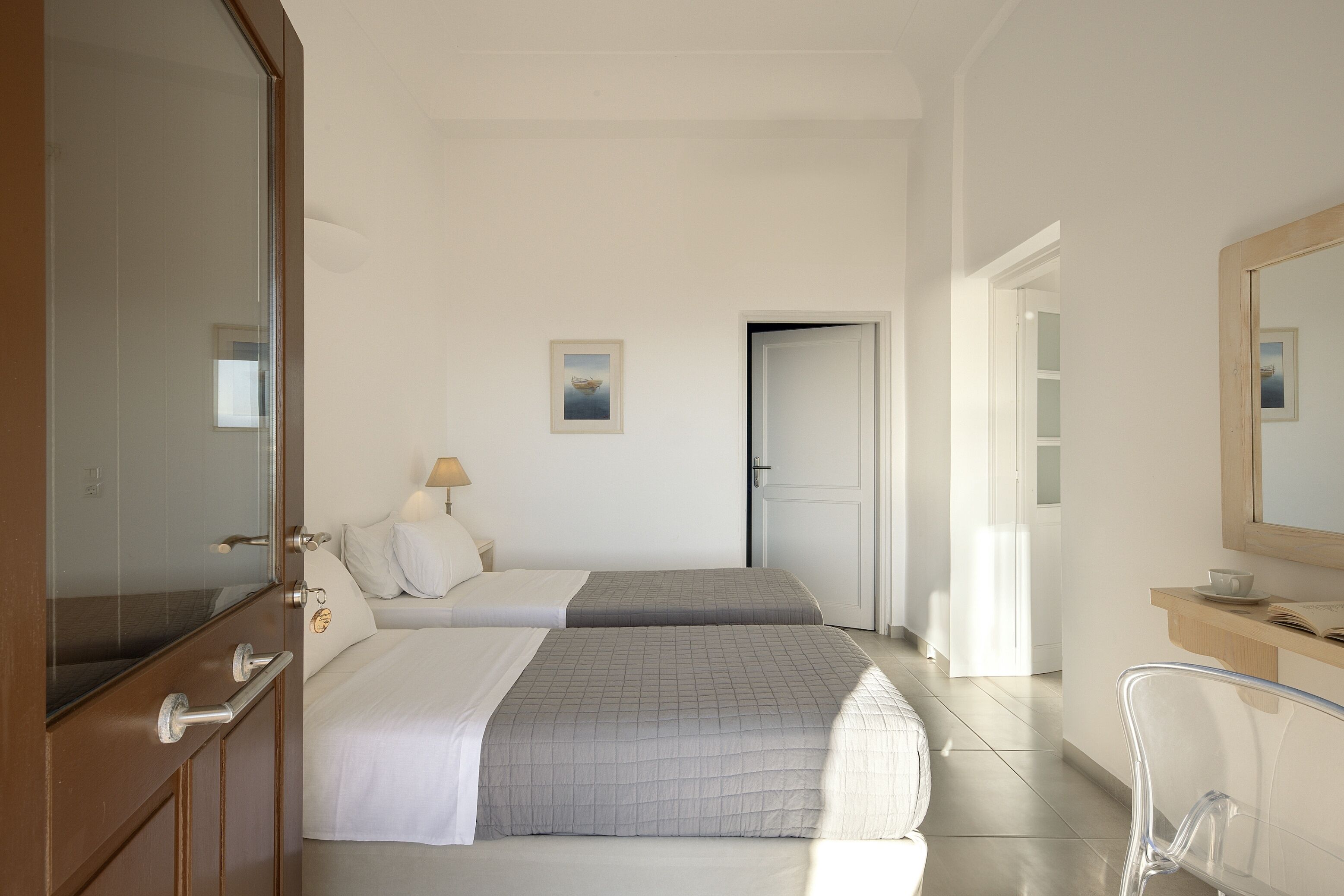 A hotel room with two beds, a wooden wardrobe, a mirror, a bedside table with a lamp, and a view of the hallway.