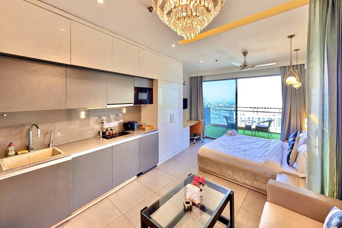 A modern hotel room with a kitchenette, a glass coffee table, a bed with a view, and a chandelier.