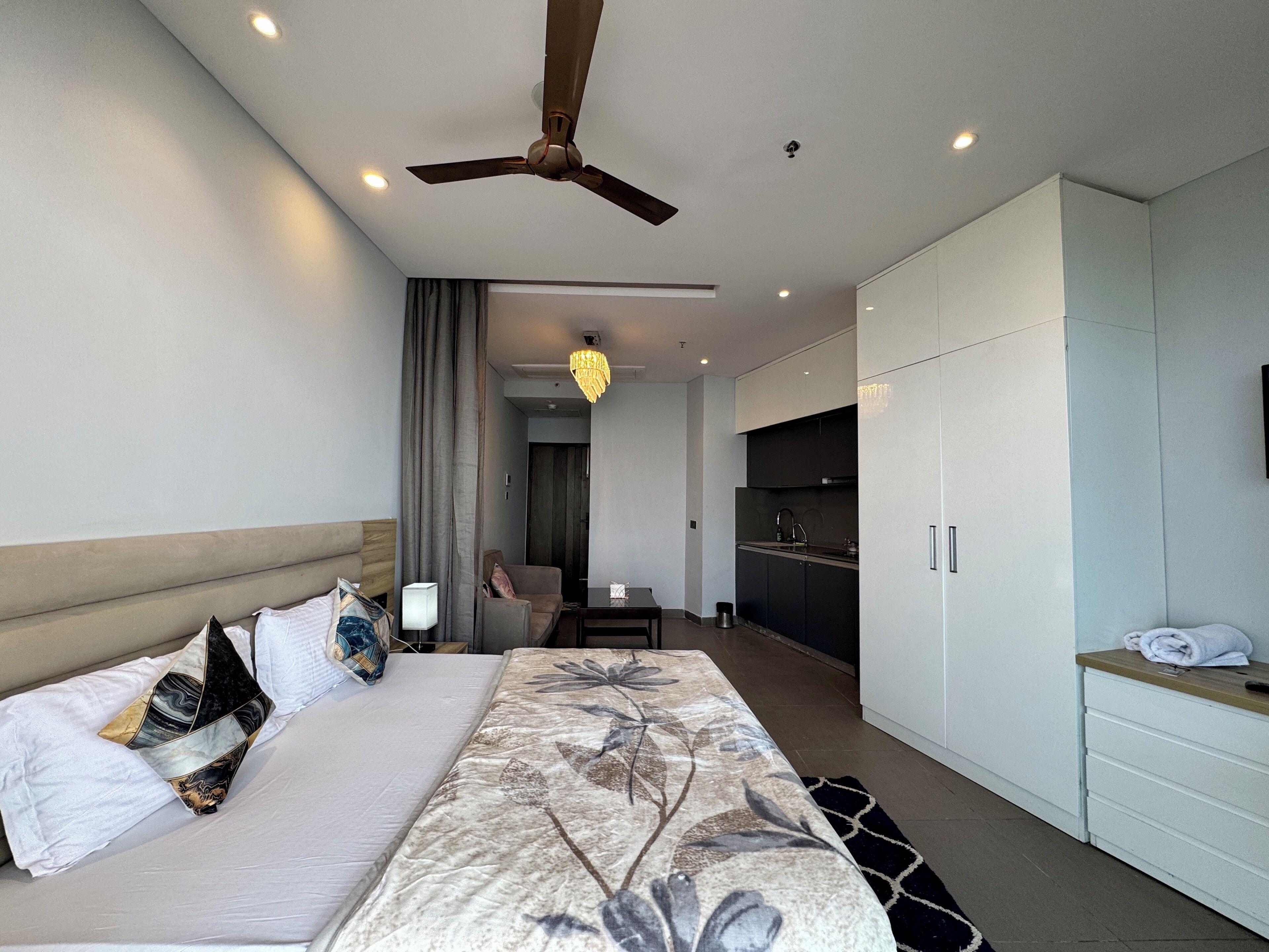 A modern hotel room with a large bed, a ceiling fan, a desk, and a kitchenette.
