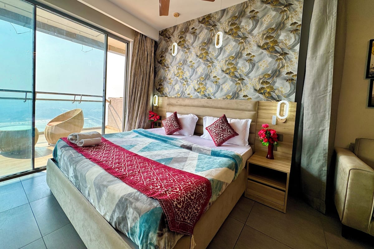 A bedroom with a large bed, a ceiling fan, a nightstand with a vase of flowers, and a view of the ocean through a sliding door.