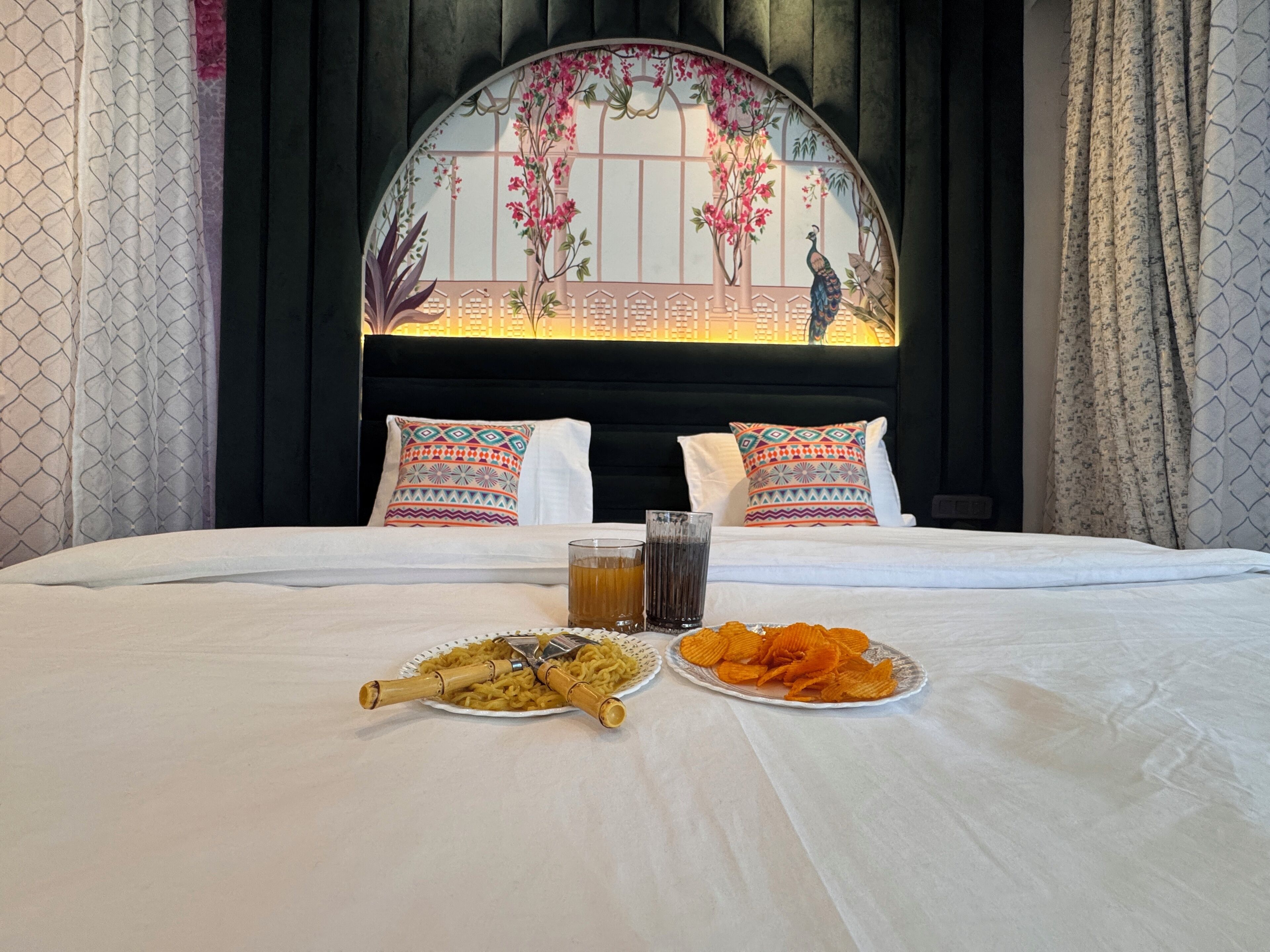 A neatly made bed with two pillows, a plate of noodles, a plate of chips, and two glasses of a beverage.