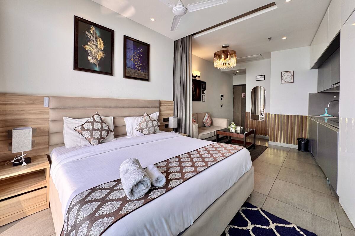 A modern hotel room with a large bed, a ceiling fan, artwork on the walls, a chandelier, and a view of the kitchen area.