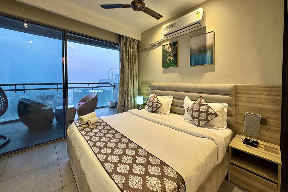 A hotel room with a large bed, a ceiling fan, an air conditioning unit, a nightstand, and a view of the city through large windows.