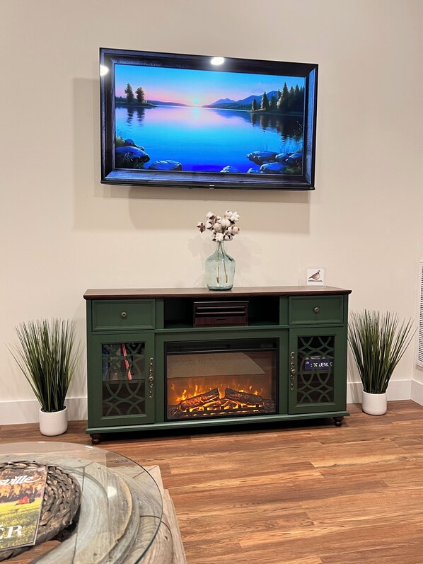 A living room with a flat-screen TV mounted on the wall, a green entertainment center with a fireplace, and two potted plants.