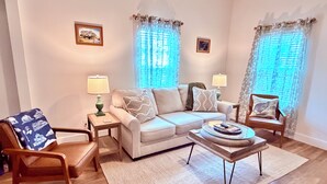 Un soggiorno con un divano, due tavolini, un tavolino da caffè, una poltrona e due finestre con tende.