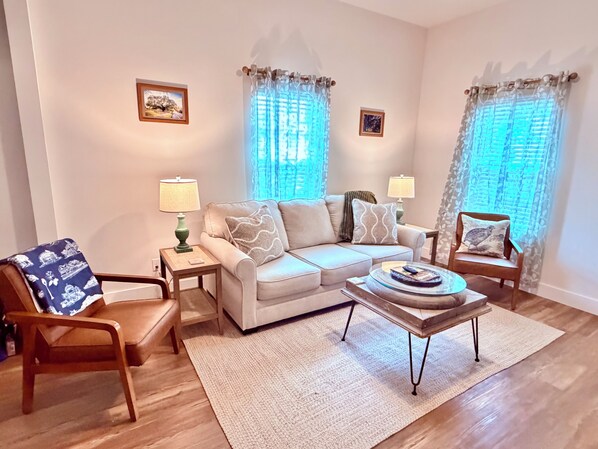 A living room with a sofa, two side tables, a coffee table, a chair, and two windows with curtains.