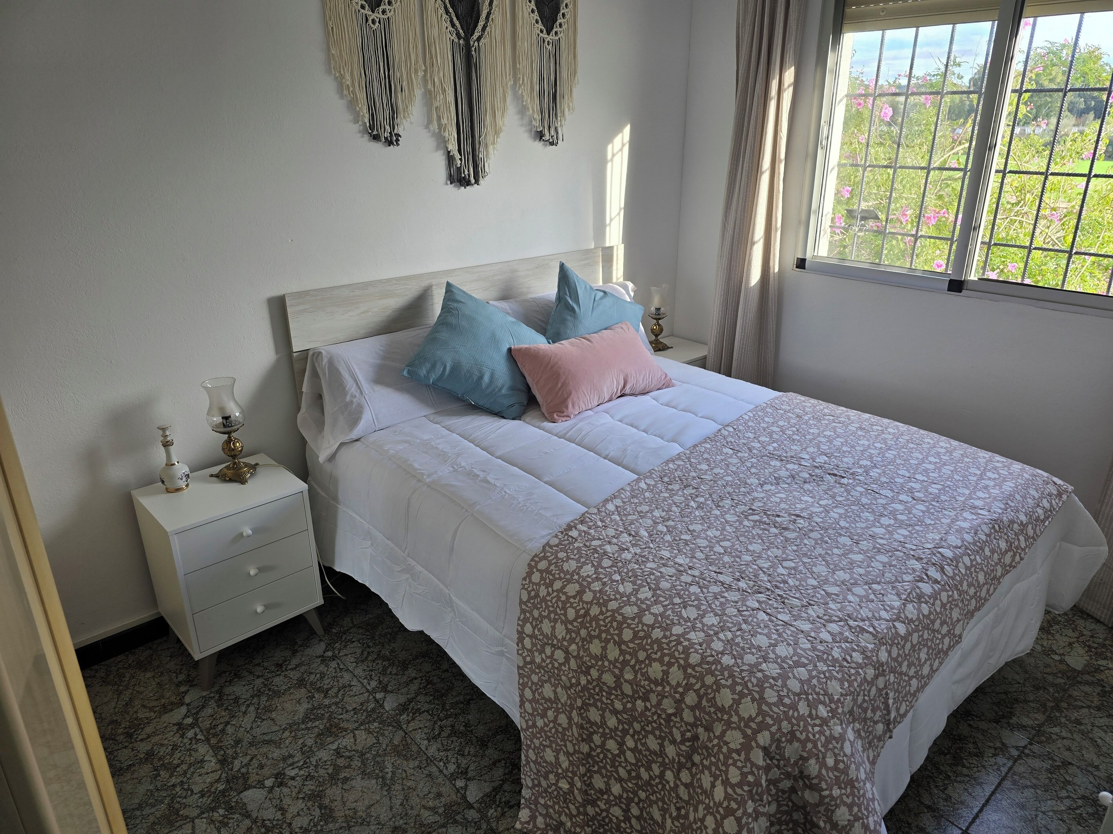 Un lit bien fait, recouvert d’une couvre-lit à motifs, une table de chevet avec une lampe et une fenêtre donnant sur un paysage verdoyant.