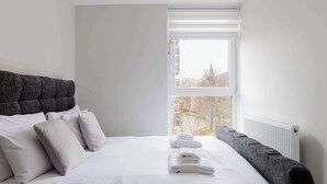 A neatly made bed with white linens and pillows, a grey upholstered headboard, and a window with blinds. - Contemporary 3BDR Retreat in South West London (Greater London)