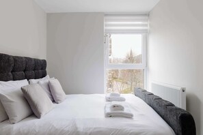 Ein ordentlich bezogenes Bett mit weißen Leinen und Kissen, ein grauer Polsterkopf und ein Fenster mit Jalousien.