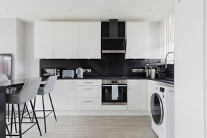 A modern kitchen with white cabinets, a black countertop, a built-in oven, a microwave, a coffee maker, and a washing machine. - Contemporary 3BDR Retreat in South West London (Greater London)