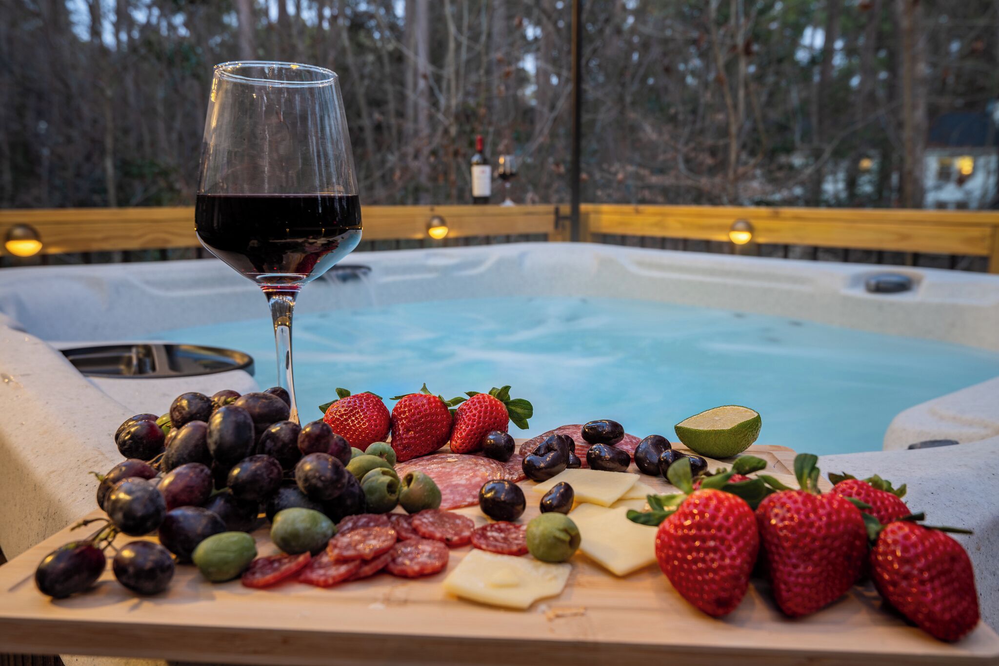A hot tub surrounded by a wooden deck with a variety of fruits and cheeses, and a glass of red wine.