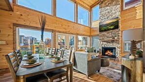 Une pièce spacieuse avec une grande table à manger en bois, dressée pour quatre personnes, une cheminée en pierre et de grandes fenêtres offrant une vue sur l’extérieur.