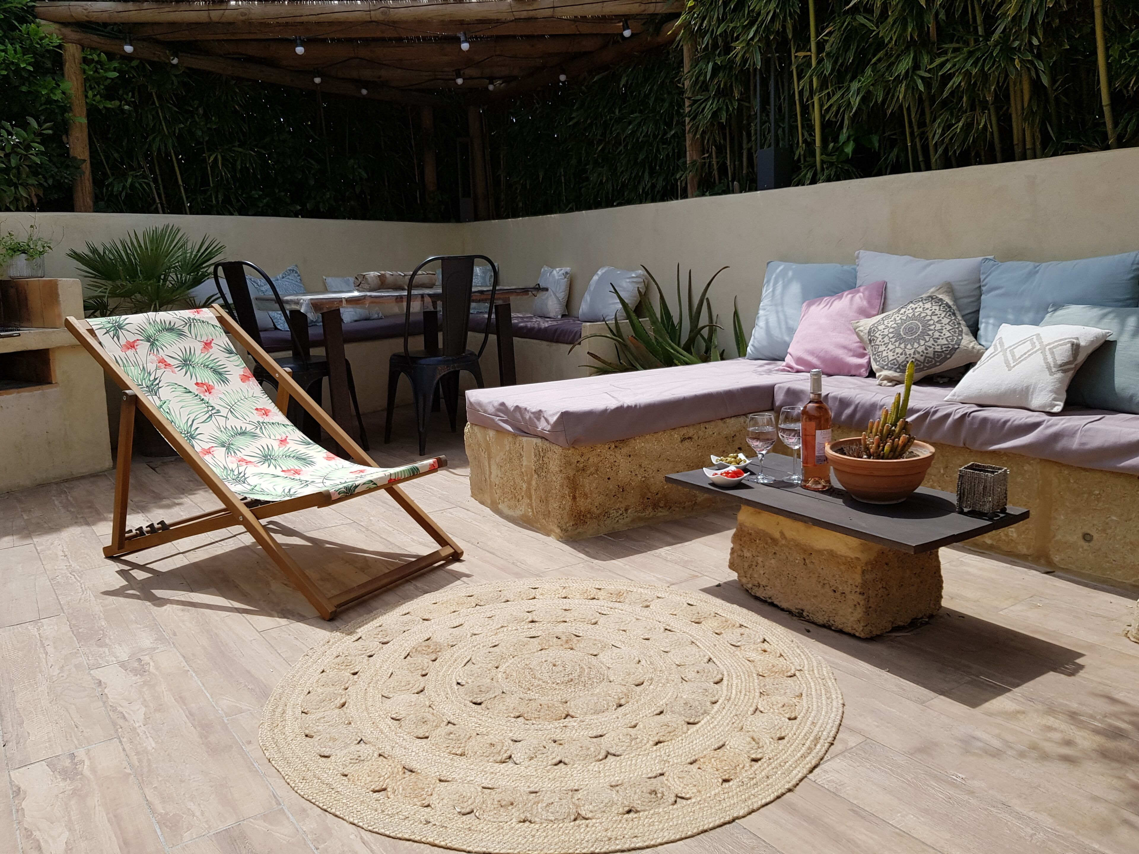An outdoor patio with a cushioned seating area, a wooden chair, a round woven rug, and a table with a potted plant and glassware.