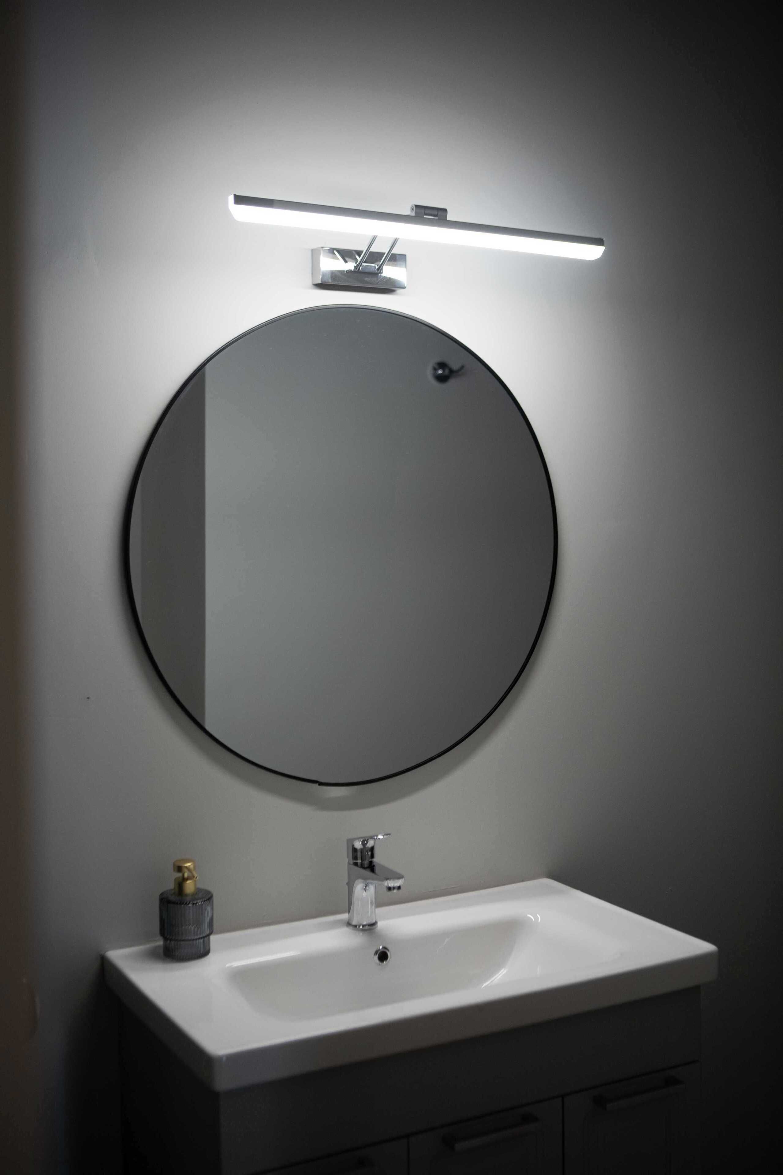 Un baño moderno con un espejo redondo, una lámpara rectangular arriba y un lavamanos blanco con grifo.