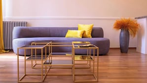 Una sala de estar moderna con un sofá morado, dos mesas auxiliares con marcos dorados y un jarrón con flores naranjas.