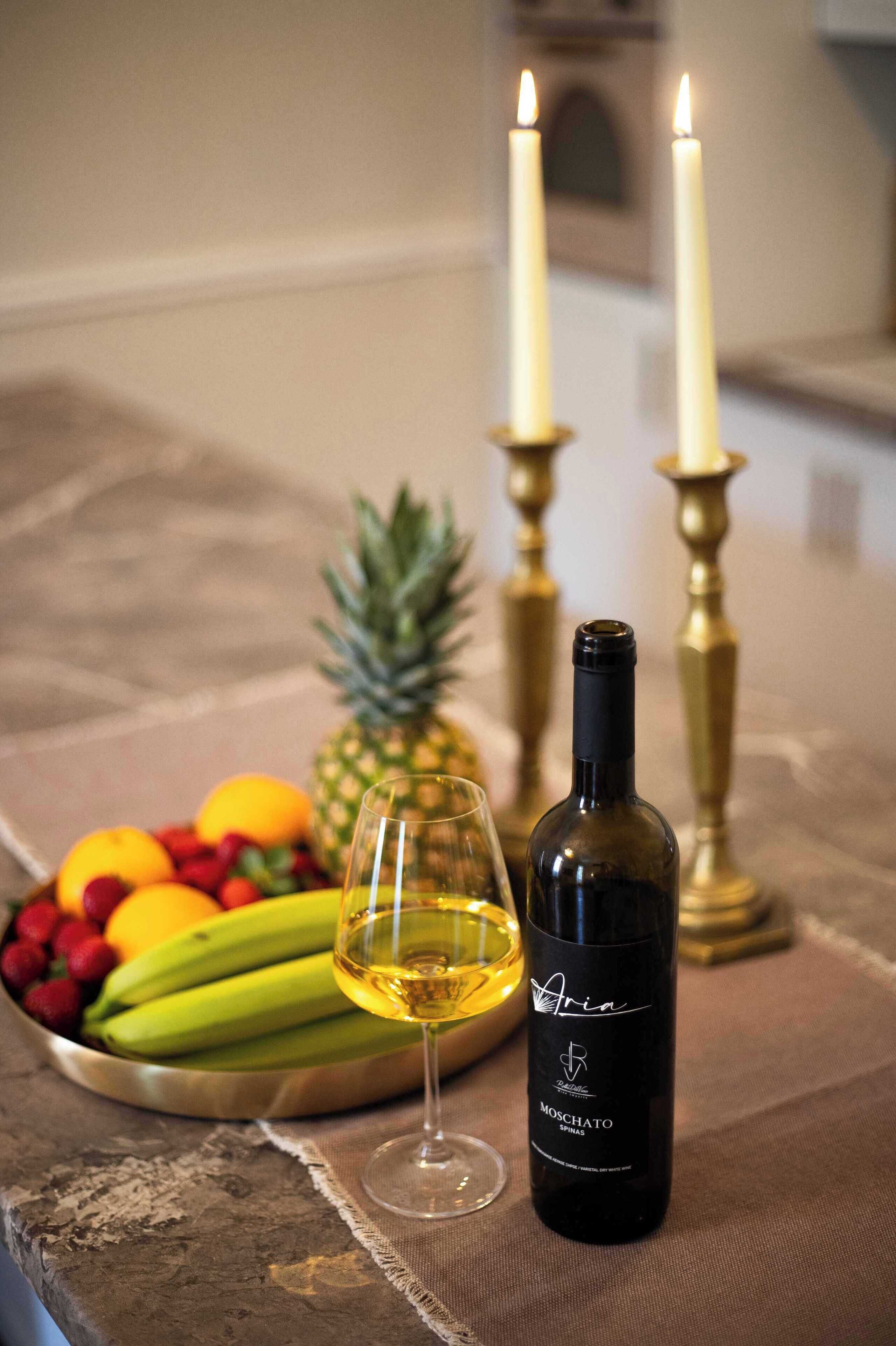 A bottle of Moschato wine, a glass of wine, and a fruit bowl with bananas, strawberries, and oranges on a table with two lit candles.