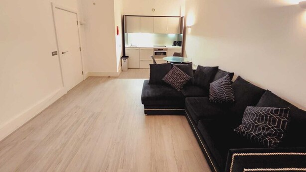 Una sala de estar moderna con un sofá seccional negro, una mesita redonda y una cocina al fondo.