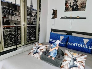 A bedroom with a bed, two blue pillows, a plush toy, a window with a view of a building, and a painting on the wall. - Room with breakfast included (Isles-lès-Villenoy)