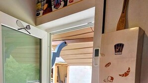 A room with a wooden ceiling, a sliding glass door leading to an outdoor balcony, a fire extinguisher mounted on the wall, and a small table with a potted plant.