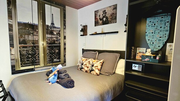 A bedroom with a bed, pillows, a stuffed toy, a window with a view of the Eiffel Tower, a shelf with items, and a wall with a picture.