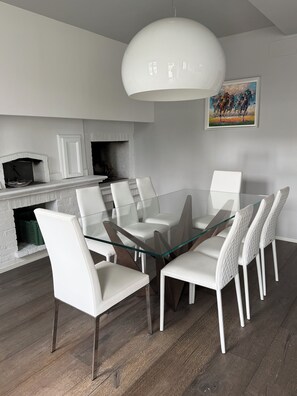 Un comedor moderno con una mesa de vidrio y sillas blancas, chimenea de ladrillo blanco y una lámpara colgante grande, también blanca.