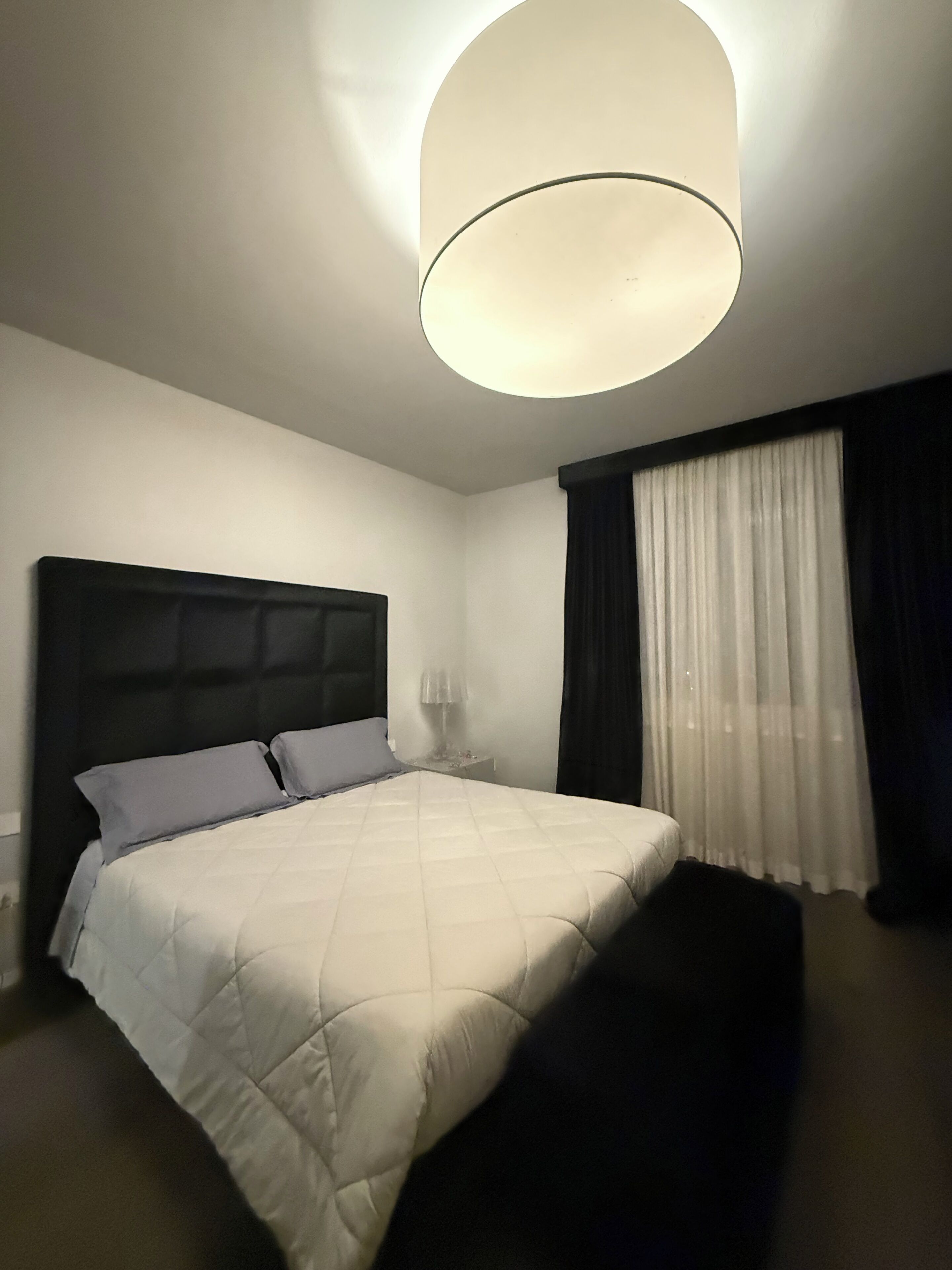 Een moderne hotelkamer met een groot bed, een hoofdbord, een nachtkastje en een groot raam met gordijnen.