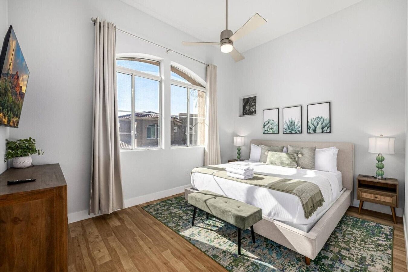 A bedroom with a large bed, a ceiling fan, a window with curtains, a bedside table with a lamp, and a small bench.