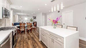 A modern kitchen with white cabinetry, a central island, and a dining area with wooden chairs and a table. - Lovely, spacious & peaceful 3-bedroom house in New Orleans w AC & outdoor space. (New Orleans)