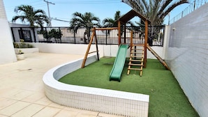 Un terrain de jeux avec un toboggan et des structures d’escalade, entouré d’un mur recouvert de carreaux blancs et d’une clôture noire.