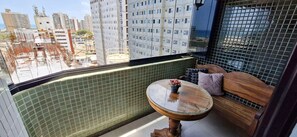 Een balkon met uitzicht op hoge gebouwen, een ronde tafel met een bloevas en een met kussens uitgeruste bank.