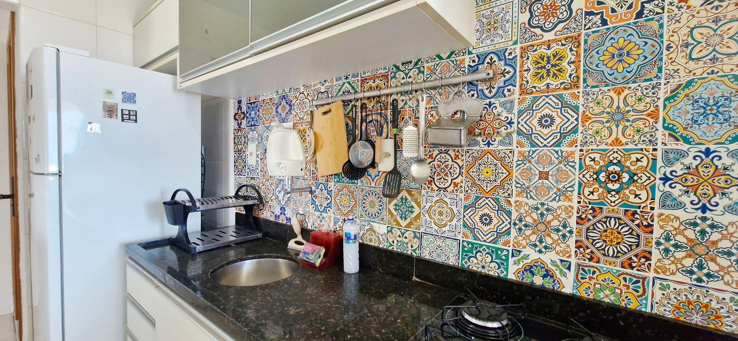A kitchen with a white refrigerator, a black countertop, and a sink. The backsplash is colorful with a patterned tile design.