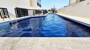 Une piscine avec un escalier en métal, entourée d’un mur carrelé et d’un bâtiment doté de grandes fenêtres.