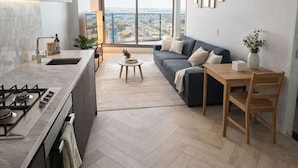 A modern kitchen with a gas stove, a dining area with a table and chairs, a sofa, and a large window offering a city view. - Skyline Sanctuary | L35 Bay & City Views (Southbank)