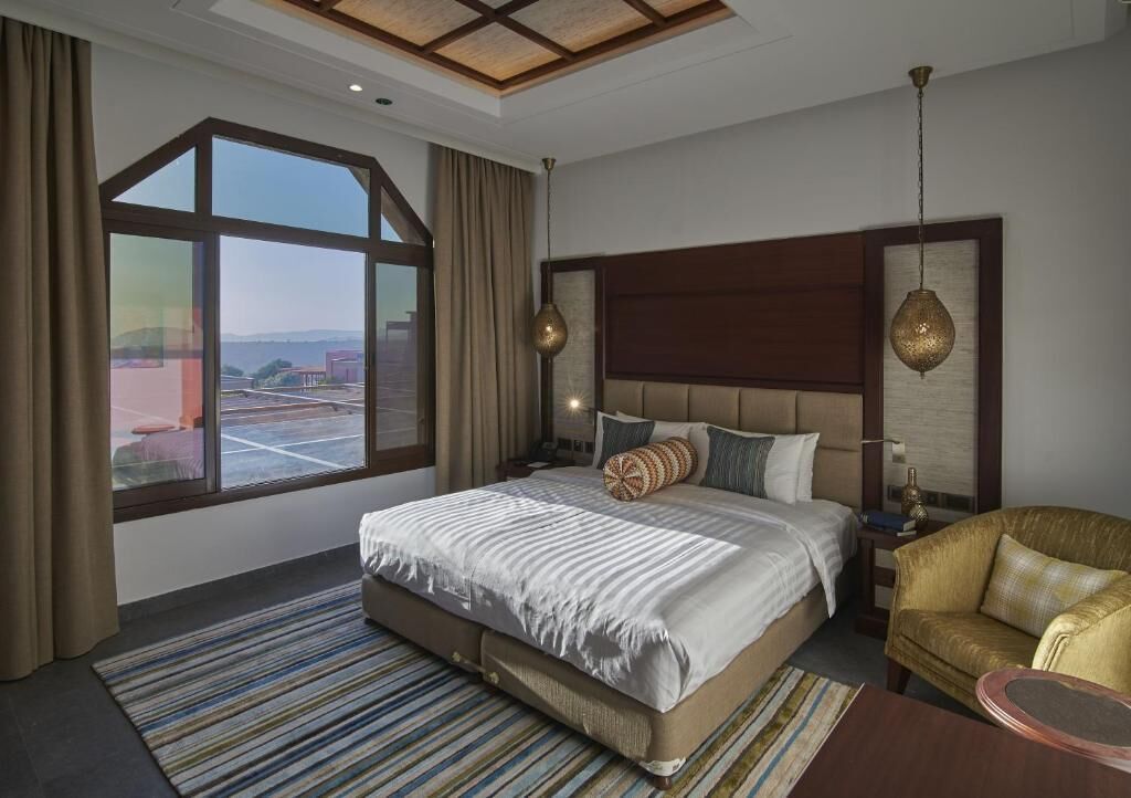Una habitación de hotel moderna con una cama grande, un sofá, una mesita y un ventanal con vistas a la ciudad.