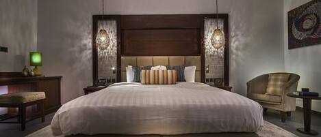 Una cama grande con cabecera de madera, dos lámparas de noche, una silla y una mesita con lámpara.
