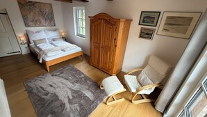 A bedroom with a bed, wooden wardrobe, two rocking chairs, a rug, and framed pictures on the wall.