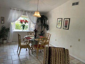 A dining area with a round glass table, wooden chairs, a hanging light fixture, and a window with curtains. - Enjoy the Daytona 500 or Bike Week w/ friends at this Port Orange 5 bed home. (Port Orange)
