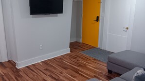 A room with a wooden floor, a grey sofa, a television mounted on the wall, and a door with a yellow accent. - Perfect 3-bedroom apartment  in  Waterbury CT (Waterbury)