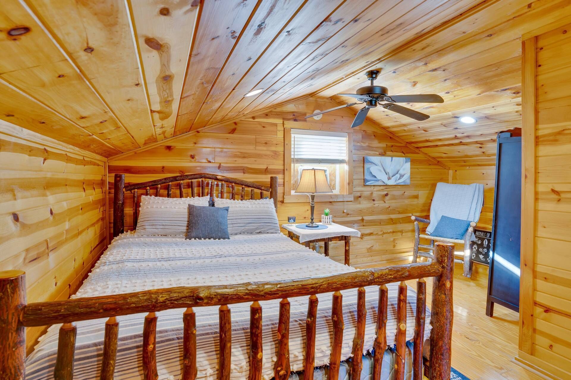 Un dormitorio de cabaña de madera con una cama, ventilador de techo, ventana, silla y refrigerador.