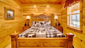 Un dormitorio de cabaña de madera con una cama, dos mesitas de noche, un ventilador de techo y una ventana con cortinas.