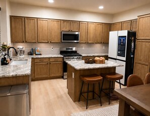 A modern kitchen with wooden cabinets, stainless steel appliances, a marble countertop, and a built-in island with stools. - Charming 3-bedroom Luxury Home, Perfect for Entertaining (Waddell)