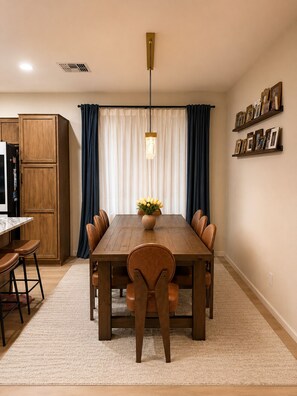 Een eetruimte met een houten tafel, stoelen, een vaas met bloemen, een hanglamp en een keuken met kasten en een koelkast.
