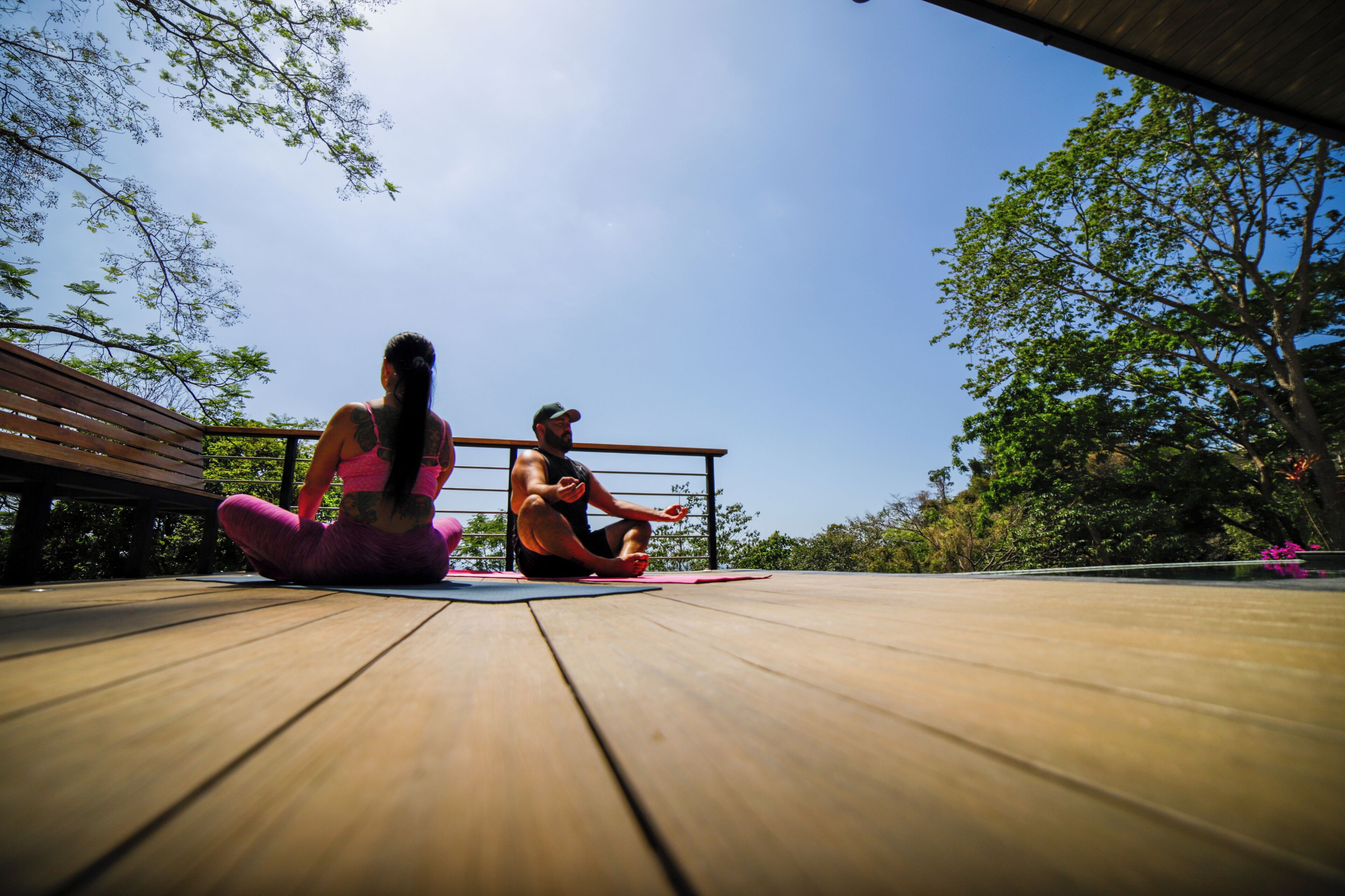 Two people sitting on yoga mats on a wooden deck with a railing and trees in the background.