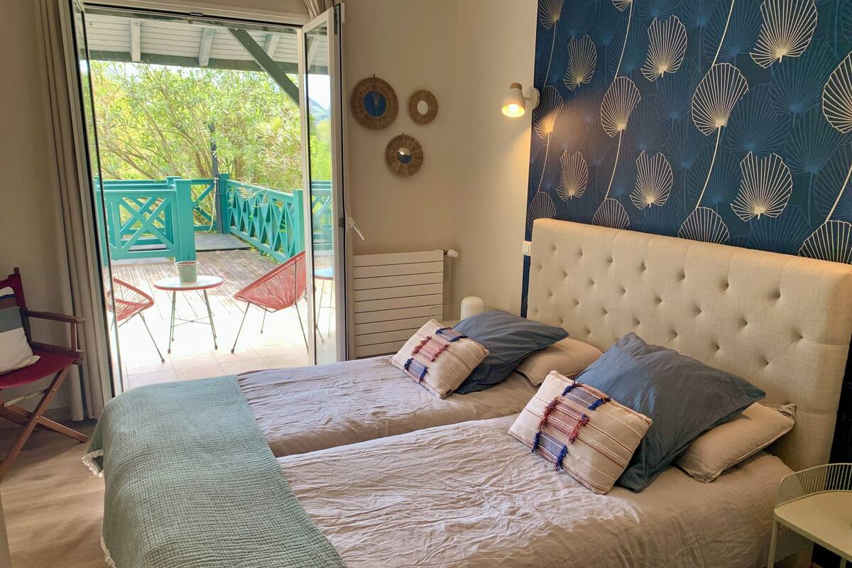 A double bed with a tufted headboard, two bedside tables, and a view of an outdoor patio with chairs and a wooden deck.