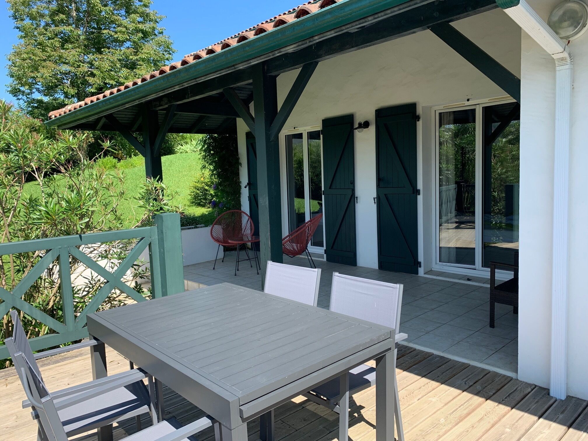 Eine Außenpatio mit Tisch und Stühlen, ein überdachter Bereich mit grünen Fensterläden und Blick ins umliegende Grün.