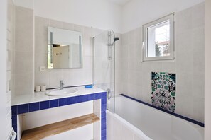 A bathroom with a white sink, blue-tiled countertop, a glass shower enclosure, and a decorative tile wall. - Villa Ongizatea (Urrugne)