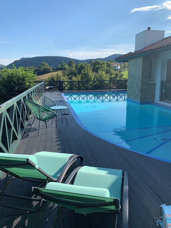 A pool area with a wooden deck, green lounge chairs, and a stone building in the background. - Villa Ongizatea (Urrugne)